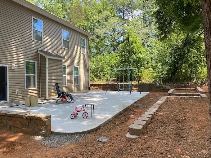 Modern house with concrete patio, children's playground equipment, and wooded backyard area.