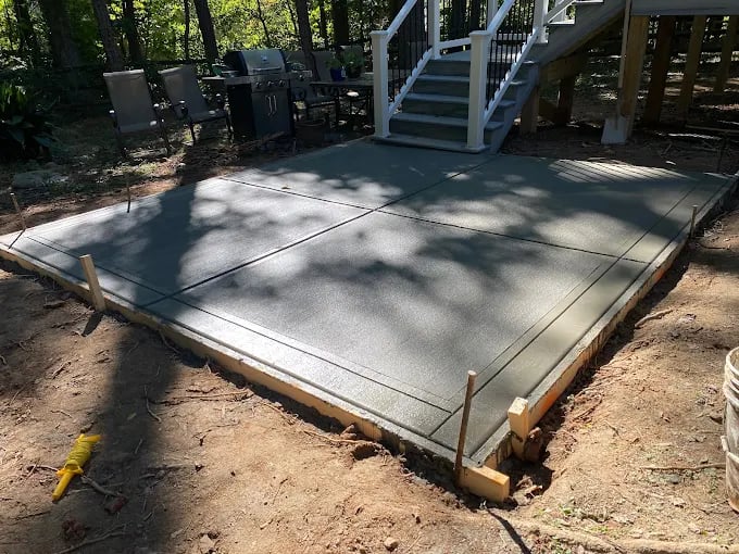 Concrete patio pad with wooden frame and railing in wooded backyard setting