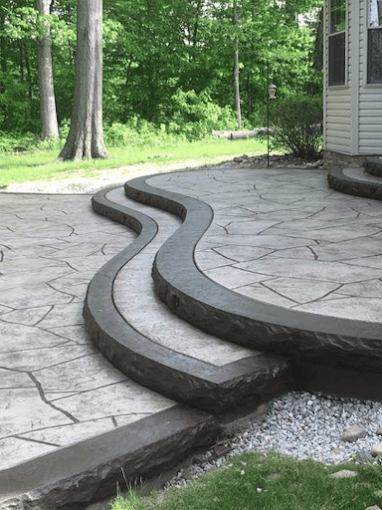 Curved stone patio with decorative border extends from house into wooded backyard